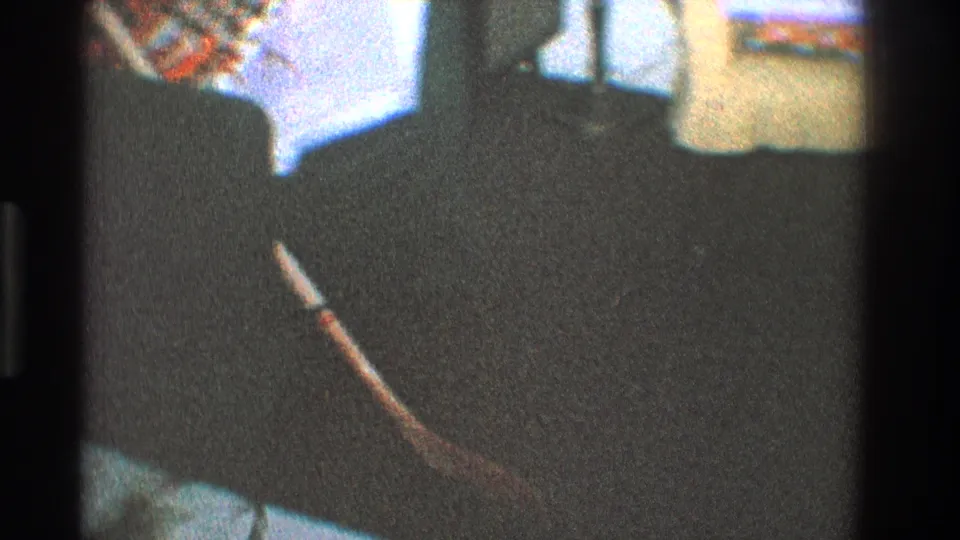 Grainy 1968 Toledo home-movie frame showing a dim living room floor with furniture silhouettes and a wrapped object near the edge.