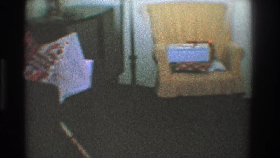 Grainy 1968 Toledo frame showing a wrapped gift on an upholstered chair near dark carpeting and adjacent furniture.