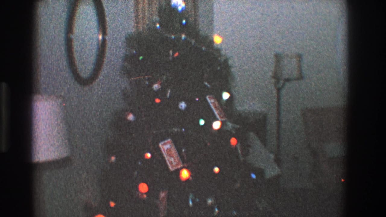1968 Toledo home-movie frame showing a Christmas tree with colored lights, ornaments, wrapped gifts, and a mirror in a dim living room.