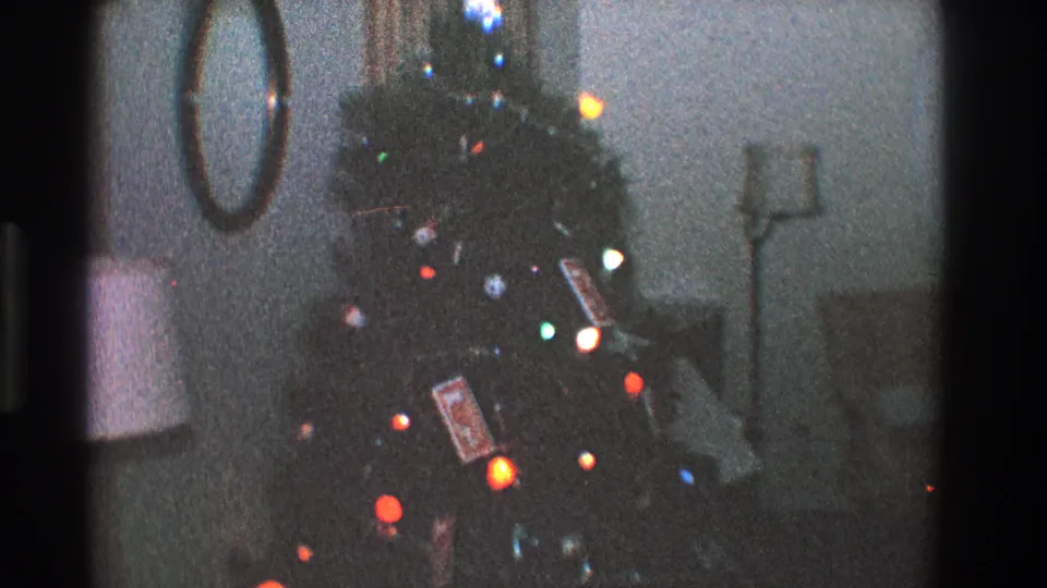 1968 Toledo home-movie frame showing a Christmas tree with colored lights, ornaments, wrapped gifts, and a mirror in a dim living room.
