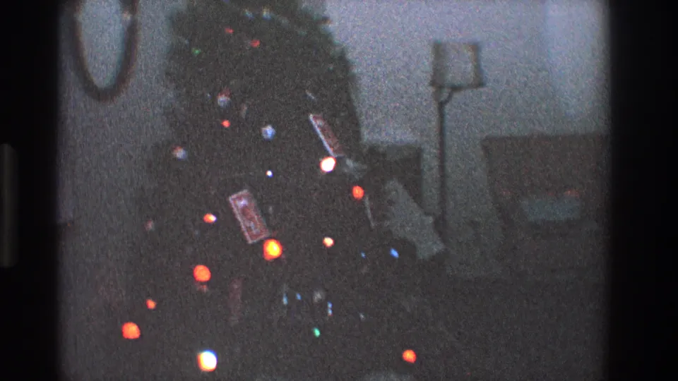 Grainy 8mm view of a Toledo living room Christmas tree, with colored bulbs, packages below, and domestic furniture behind it.