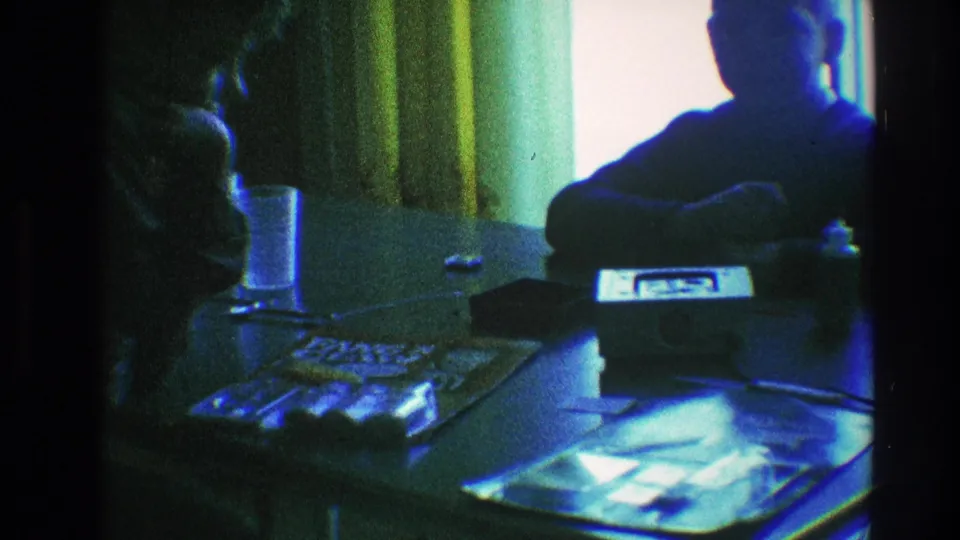 The home-movie frame shows the backlit seated figure and reflective glass table in a Toledo interior from 1968.