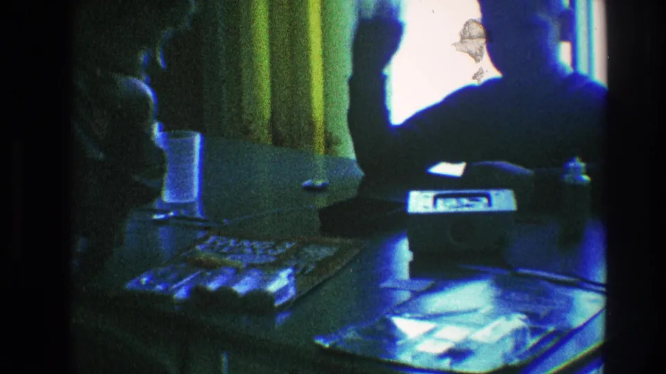 In this 1968 Toledo home-movie frame, the seated figure raises an arm near the curtained window and tabletop objects.
