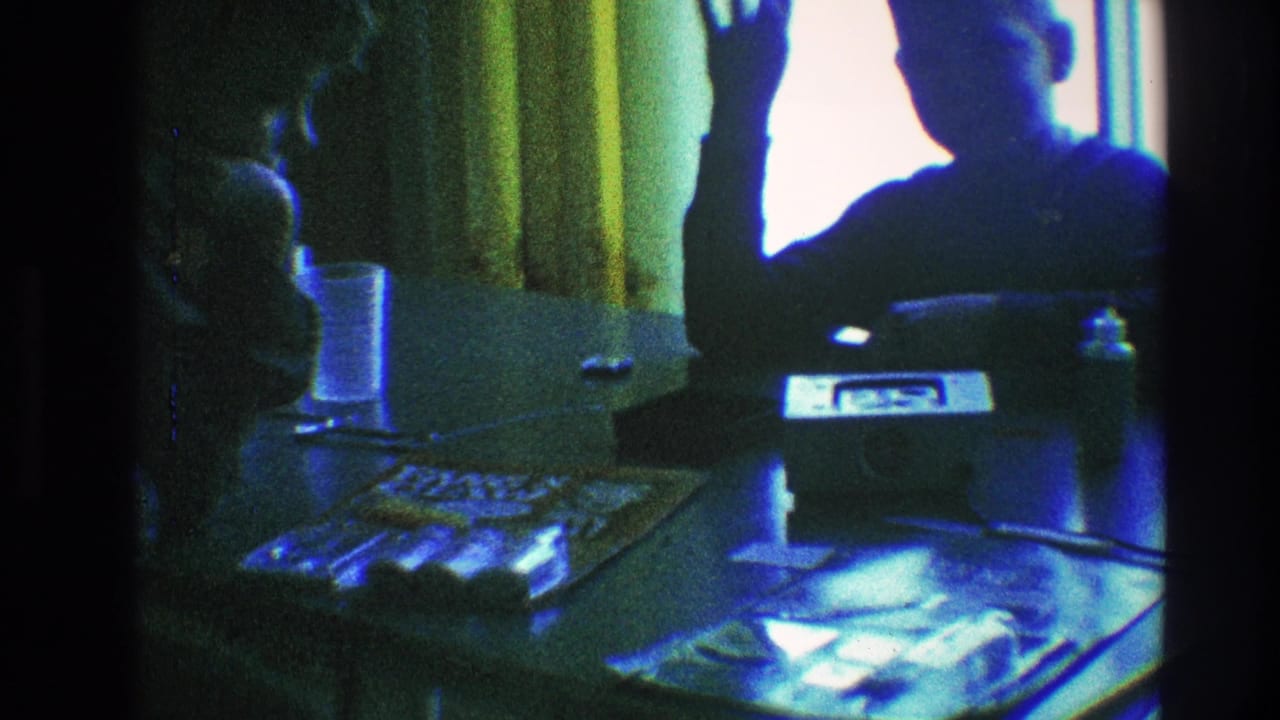 Two silhouetted people sit at a table near yellow curtains as one raises a hand toward the camera.
