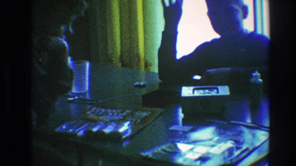 In this 1968 Toledo home-movie frame, two figures sit by a table and bright window, with one person raising a hand near yellow curtains.
