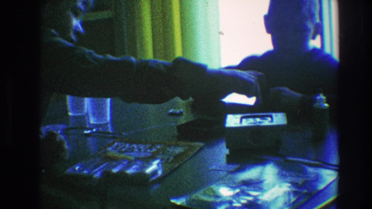 Two figures at a table with a small radio, papers, glasses, and curtains behind them.