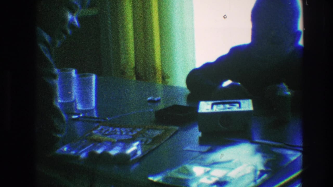 Younger figure sits at the table beside a small radio, glasses, papers, and a curtained window.