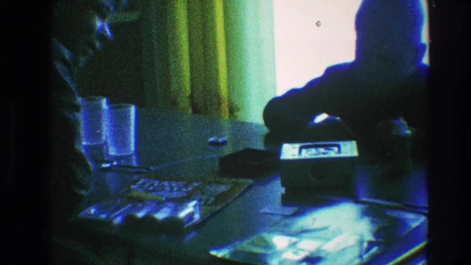 The younger boy remains near the small radio on a table in a dim Toledo interior, with glasses and papers visible under blue-green film color shifts.