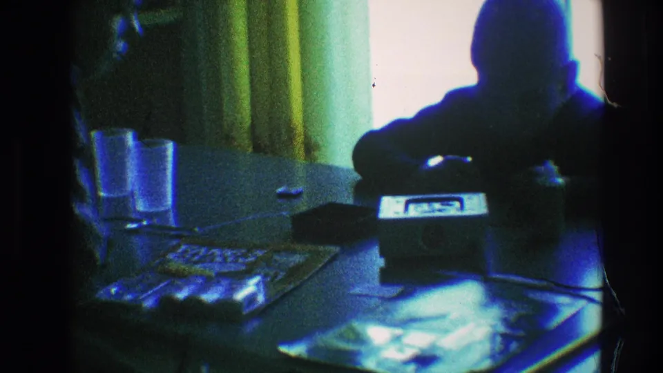 A boy leans in toward the tabletop radio during the 1968 Toledo home-movie scene, surrounded by papers, glasses, and dim interior light.
