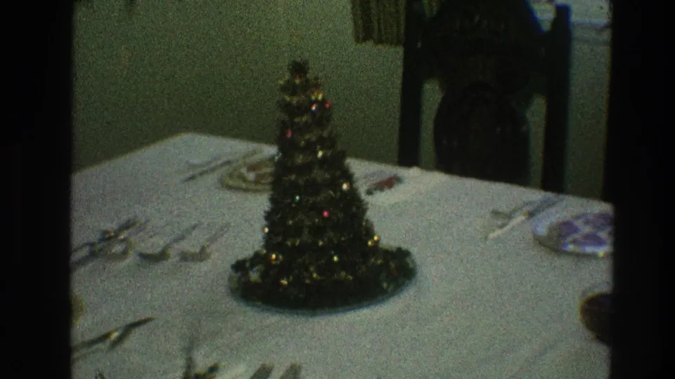 1968 Toledo home-movie frame showing a small lit tabletop Christmas tree centerpiece on a white tablecloth with plates and silverware nearby.