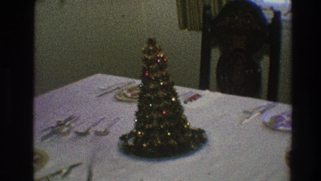 Mini Christmas tree with tinsel and colored ornaments sits on a set dining table in a Toledo, Ohio home-movie interior.