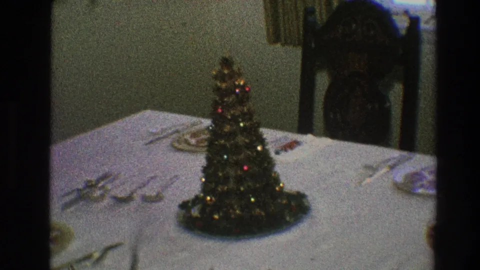 Mini Christmas tree with tinsel and colored ornaments sits on a set dining table in a Toledo, Ohio home-movie interior.