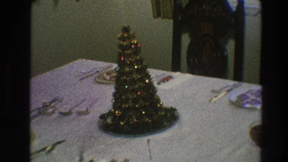 1968 home-movie still of a miniature Christmas tree centerpiece on a white tablecloth with place settings and chairs visible.