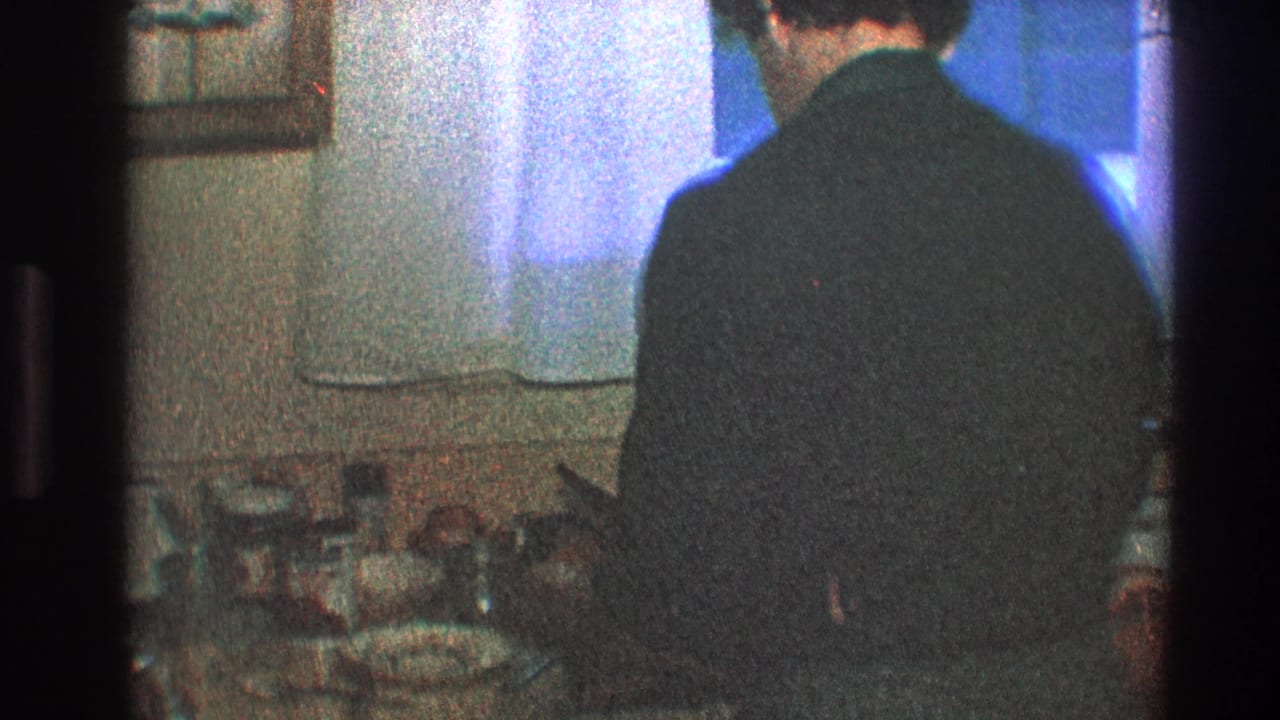Man stands near the counter with white curtains and kitchen items behind him.