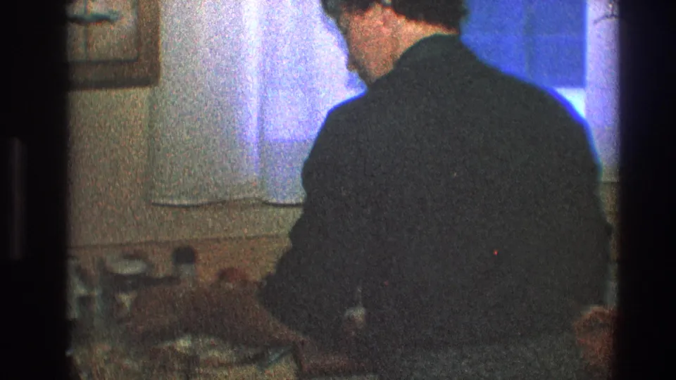 The man leans forward at the kitchen counter, suggesting food preparation or conversation during a 1968 home gathering.