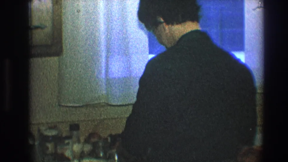 The man stands near the curtained window and counter in a private Toledo kitchen, framed softly by 8mm home-movie exposure.