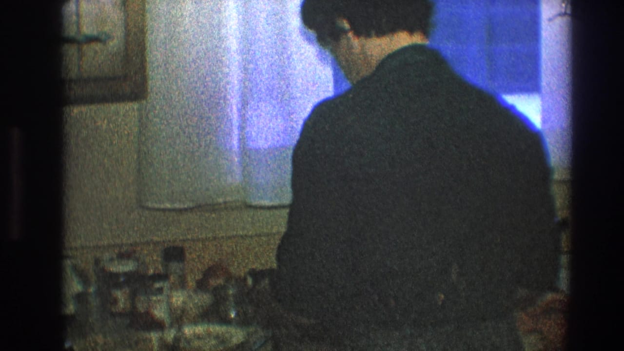 Man seen from behind at a kitchen sink beneath a curtained window.