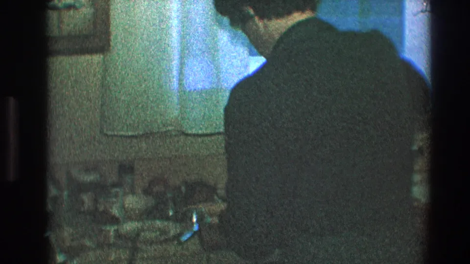 A grainy 1968 Toledo frame shows the man at the sink, back turned, with blue-green window light and a sheer curtain behind him.