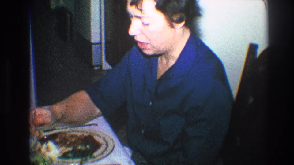 Older woman in a blue collared shirt eats dinner at a table in 1968 Toledo home-movie footage.