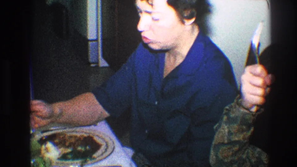 Older woman eating at a Toledo dinner table as another person's hand enters the frame in 1968 home-movie footage.