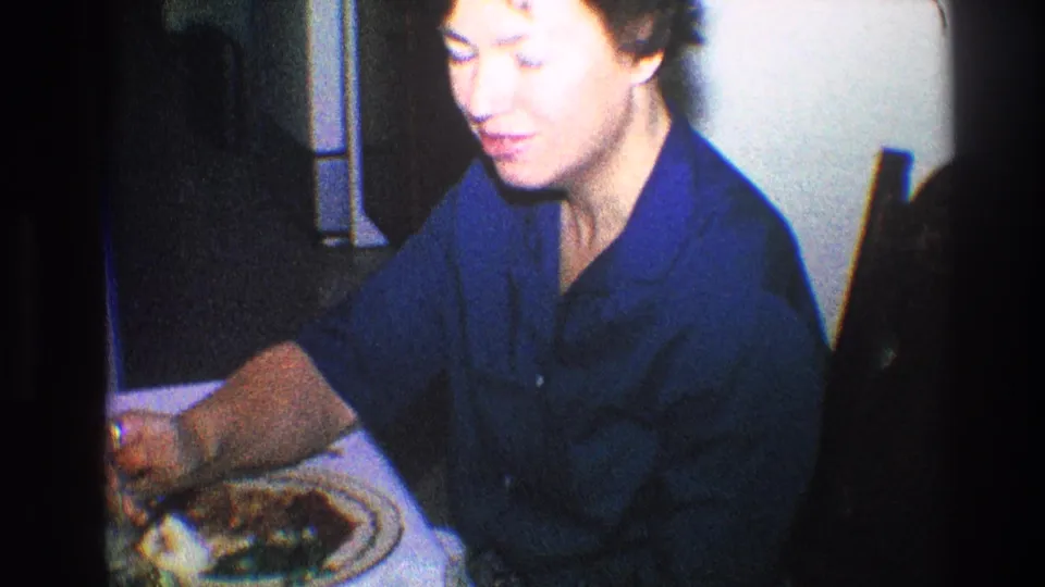 Older woman in a blue shirt begins to smile while seated at a dinner table in 1968 Toledo home-movie footage.