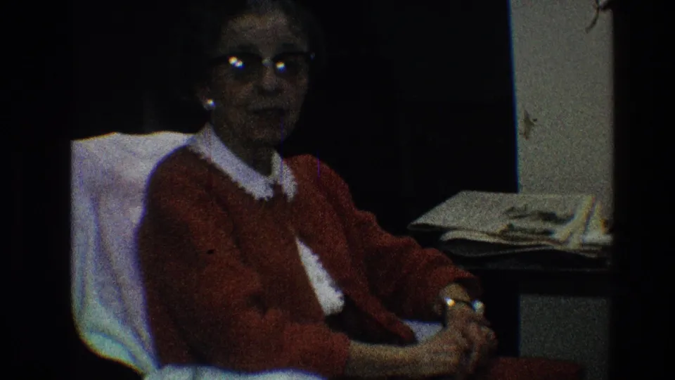 In the 1968 Toledo home movie, an older woman in glasses and a red cardigan sits in a white chair beside papers in a dim room.