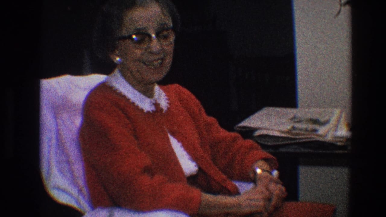 Older woman in a red cardigan sits indoors with hands clasped.