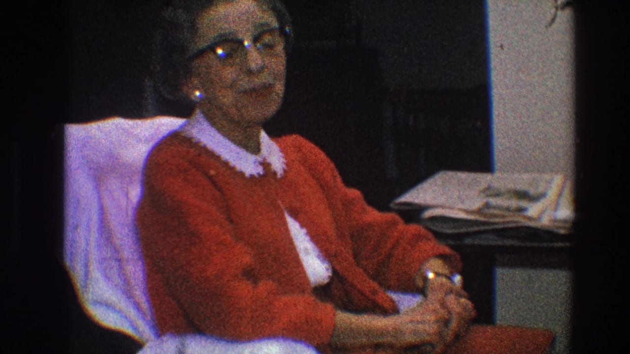 Older woman in glasses sits beside a table with papers.