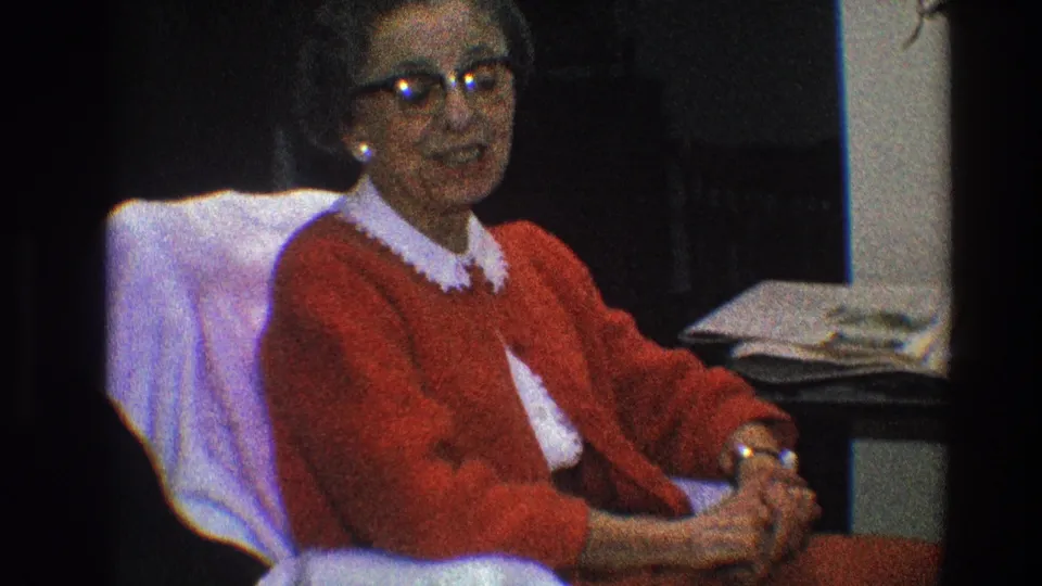 Older woman wearing a red cardigan and white collar sits with folded hands in a private Toledo interior.