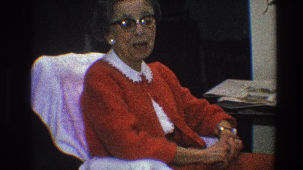 Woman in a red cardigan sits in a pale chair with hands clasped.