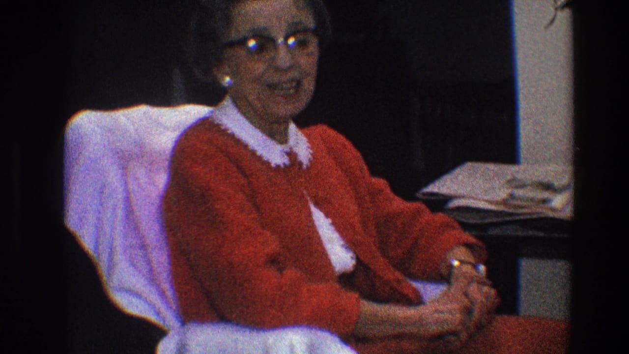 Woman in glasses sits in a chair, wearing a red cardigan and white collar.
