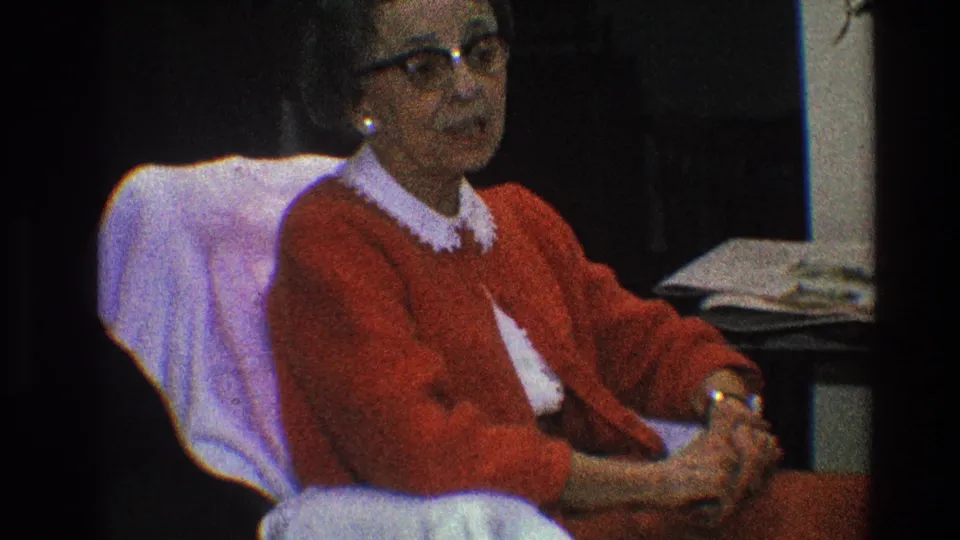 The woman is shown more in profile, seated indoors in a red cardigan with papers or books visible beside her.