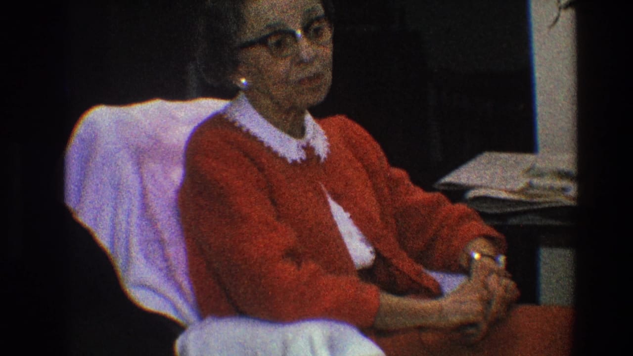Elderly woman in a red cardigan sits in a covered chair with hands clasped.