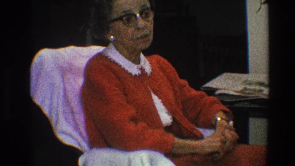 The older woman remains seated indoors in Toledo 1968 footage, wearing a red cardigan and dark-rimmed glasses.