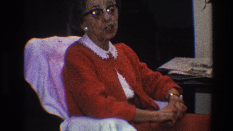 In the final sampled frame, the elderly woman appears mid-sentence in a dark domestic interior from the 1968 Toledo clip.