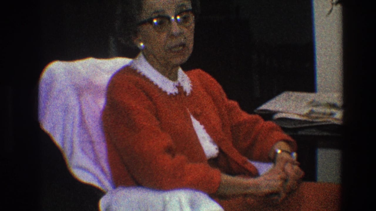 Older woman in a red cardigan sits in a white-covered chair inside a decorated room.