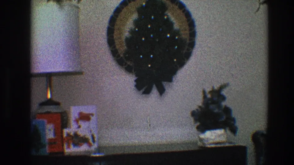 1968 Toledo home-movie frame showing a Christmas wreath on a wall above a lamp, holiday card, red box, and small plant.
