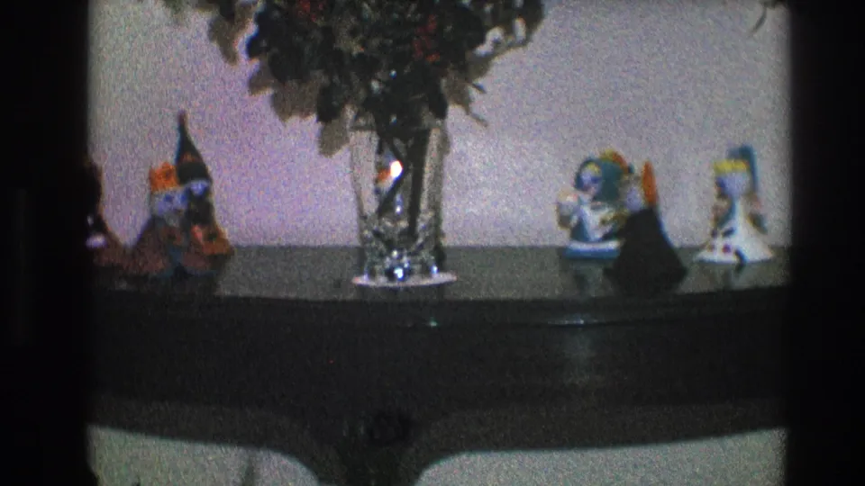 Grainy 1968 Toledo home-movie frame of a dark wood console table with a vase, greenery, and small decorative figures.