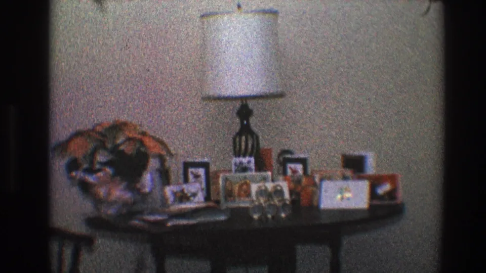 1968 Toledo interior frame with a table holding a lamp, framed photographs, floral decor, and small personal items.