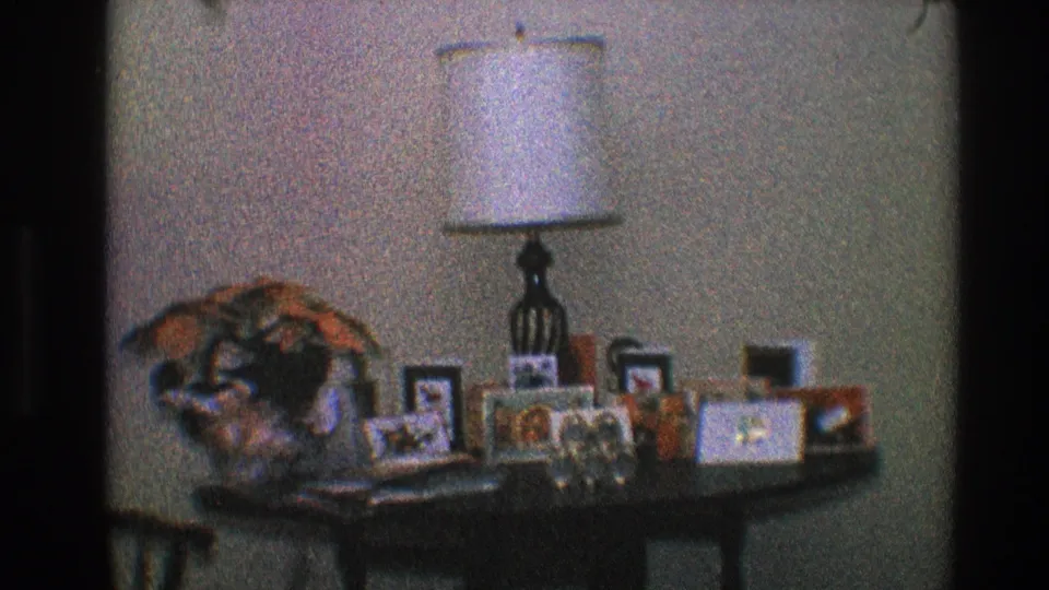 Home-movie view of a living-room table in Toledo with a lamp, framed photos, flowers, and personal keepsakes.