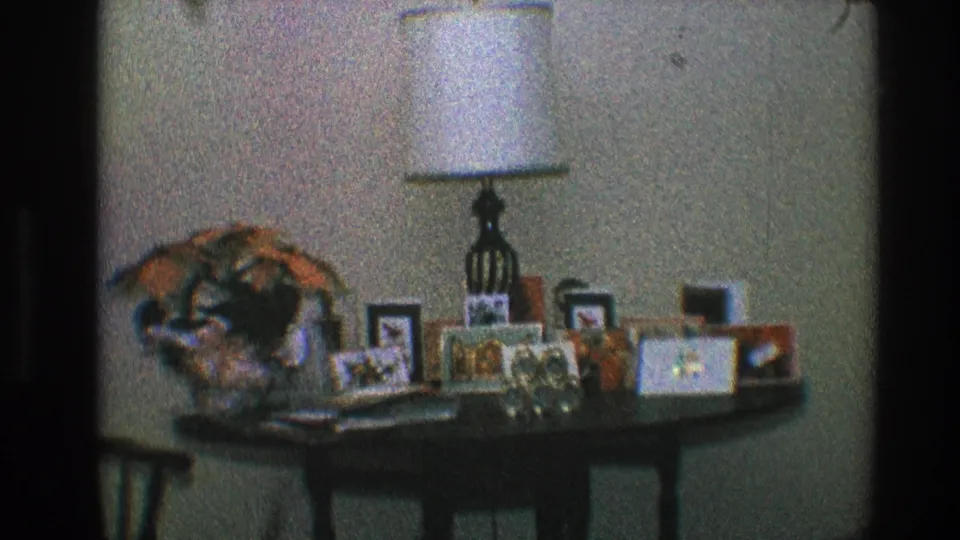 1968 home-movie still from Toledo showing a dark table, white lamp, framed pictures, and personal decor in a private room.