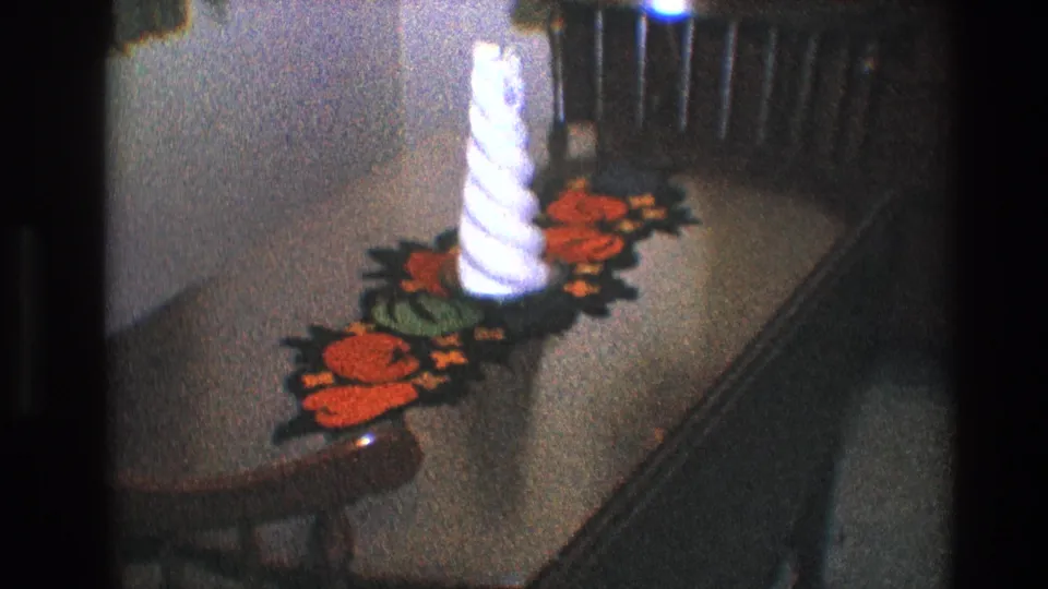 Final 1968 Toledo frame of the white spiral candle and red-green table runner on a brown dinette surface in a dim room.