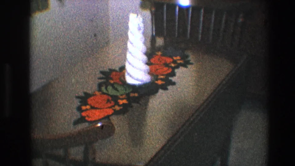 1968 Toledo home-movie frame showing a white spiral candle on a crocheted orange-and-green runner atop a wooden table.