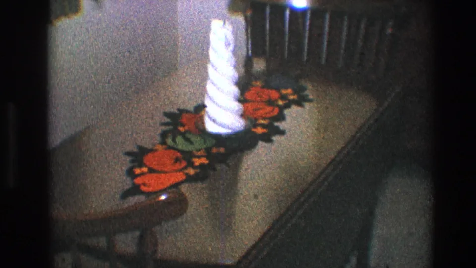 Late-1960s Toledo home-movie view of a white candle standing on a colorful crocheted runner across a wooden table.