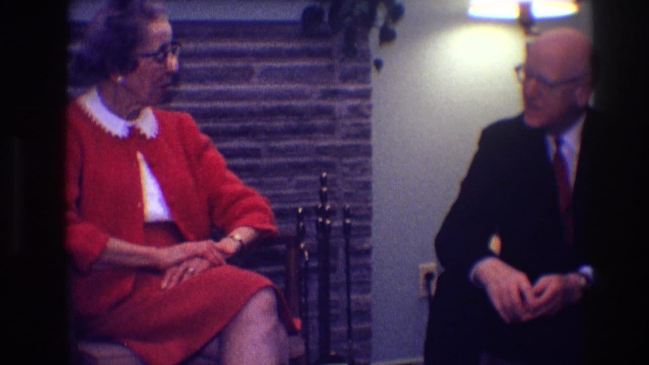Woman in a red suit faces a man in a dark suit while seated indoors.