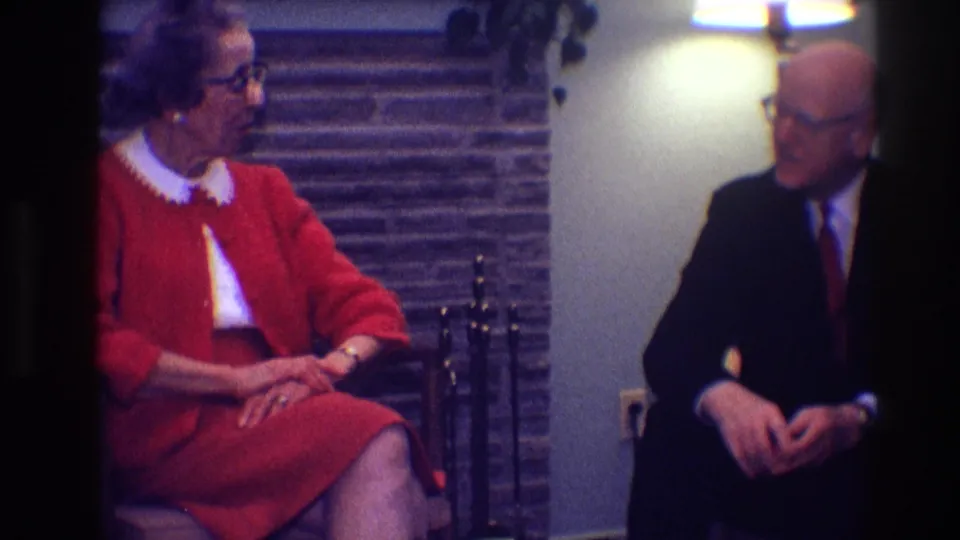 The woman in a red suit and the man in a dark suit appear to talk while seated in a Toledo living-room interior.