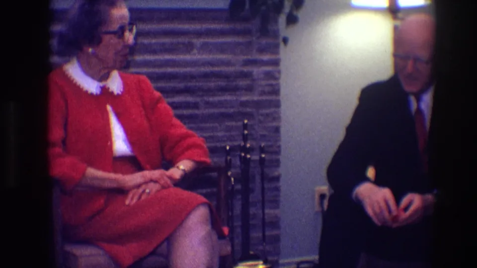 A woman in red sits with hands folded while a man in a dark suit sits nearby in the softly exposed 8mm frame.