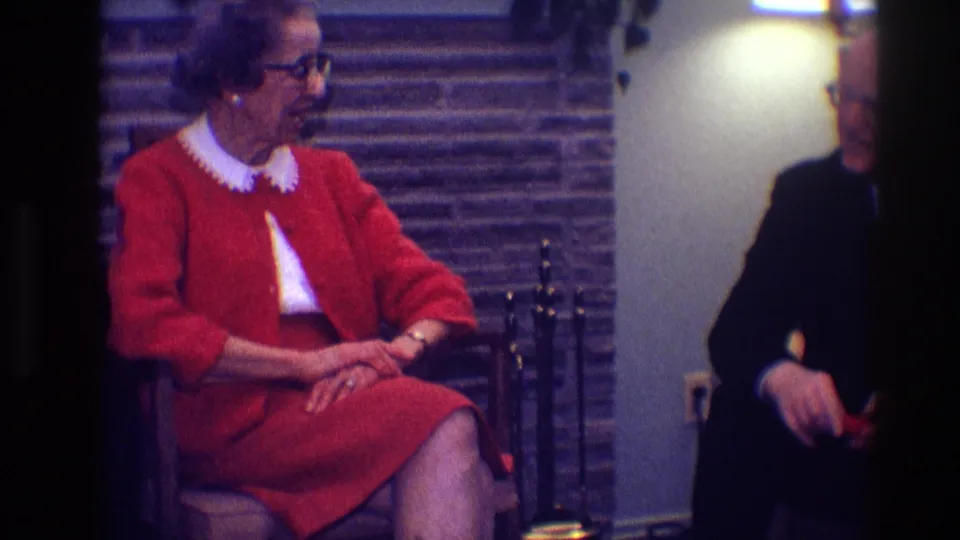 The woman in red remains seated near the fireplace tools as the man in a dark suit is partly cropped at the right side of the frame.