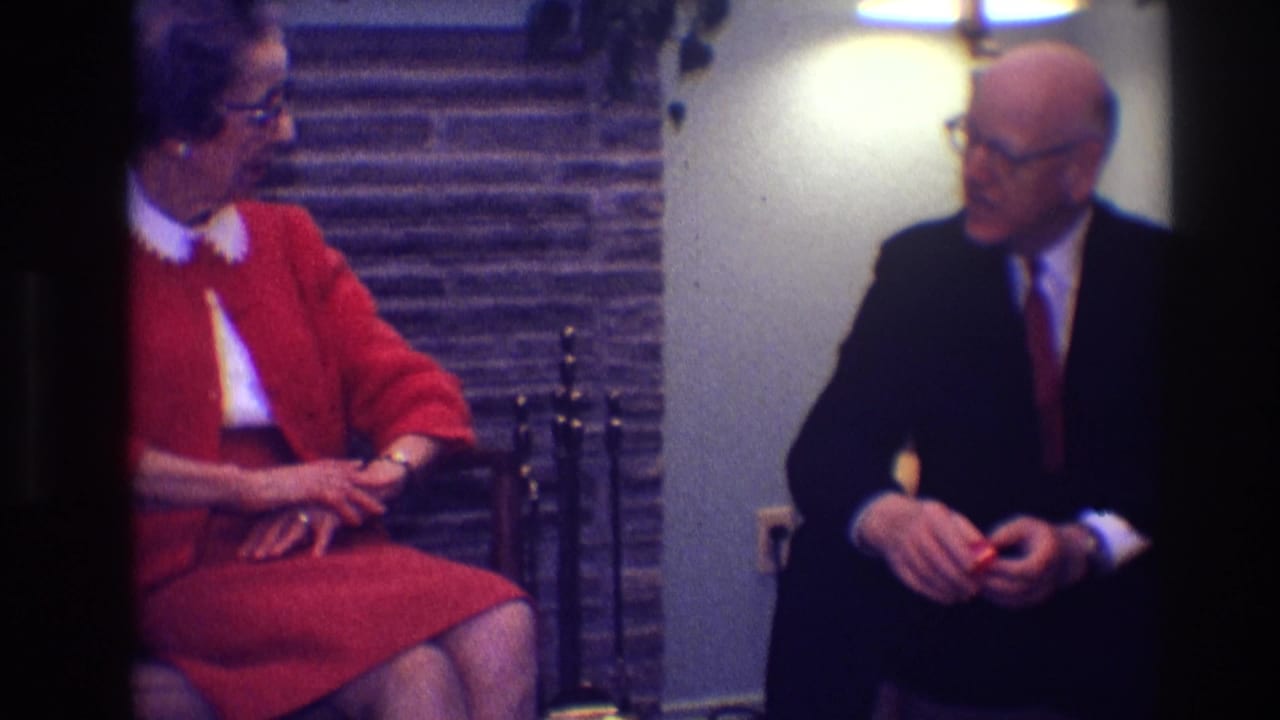 Woman in red turns toward a man in a dark suit seated nearby.