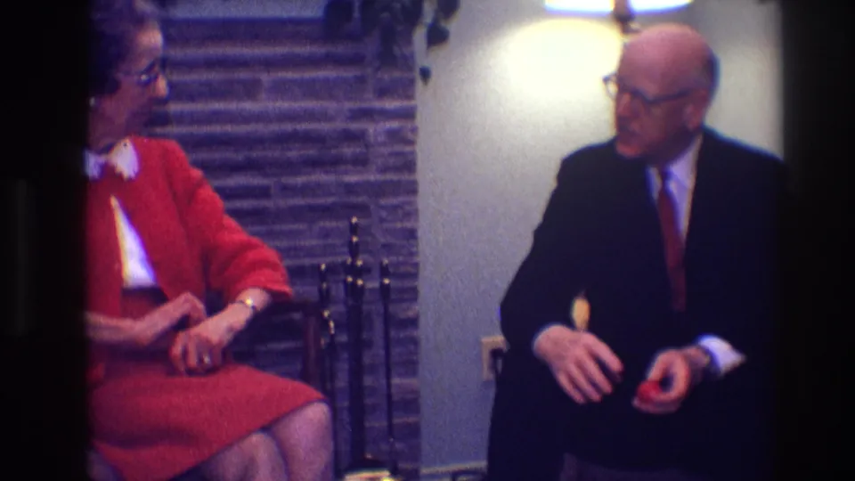 A suited older man sits near the woman in red, holding a small red object in a private Toledo interior.